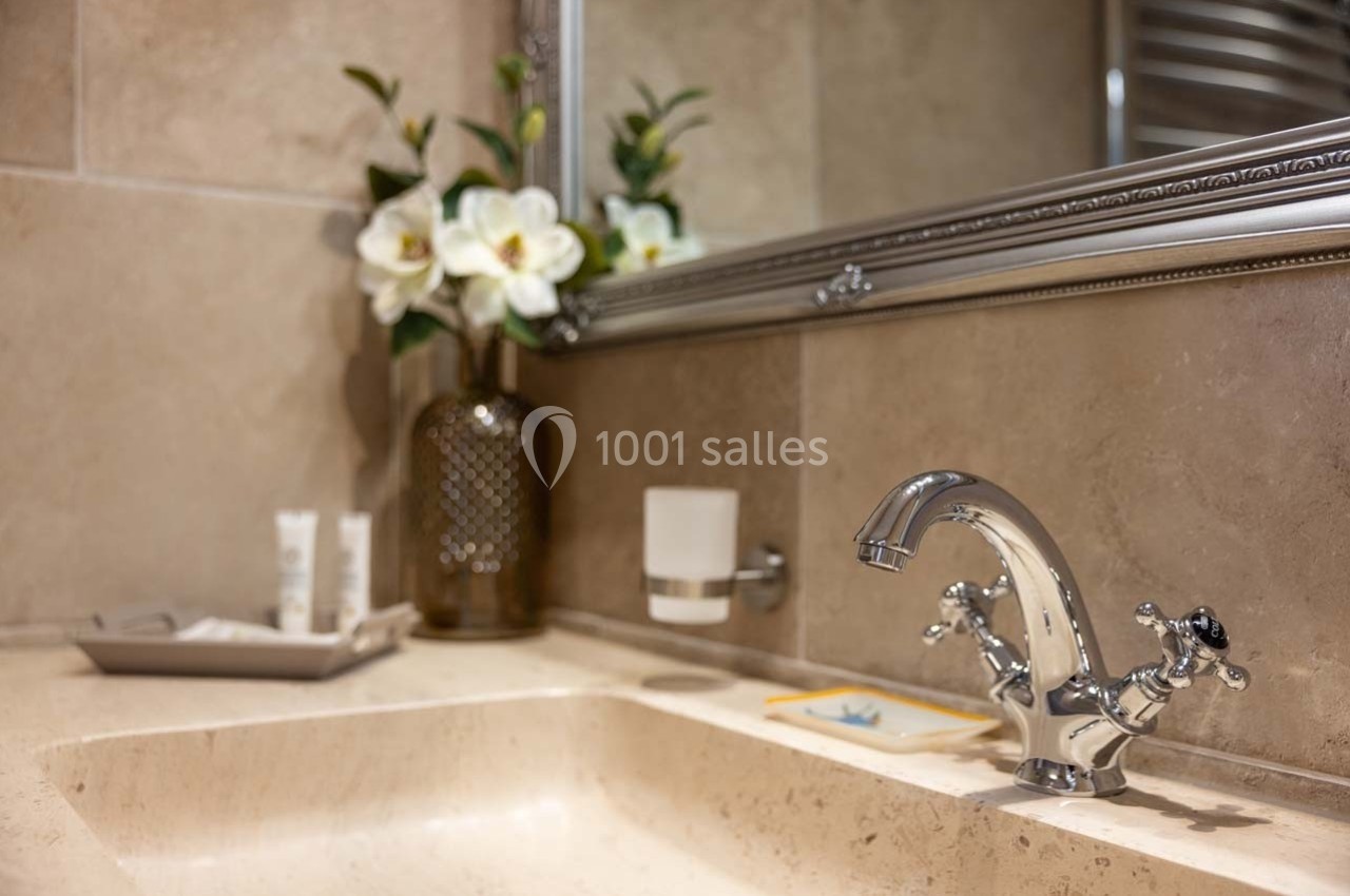 Lavabo en pierre beige avec robinet chromé, miroir encadré, vase de fleurs blanches et accessoires de toilette.