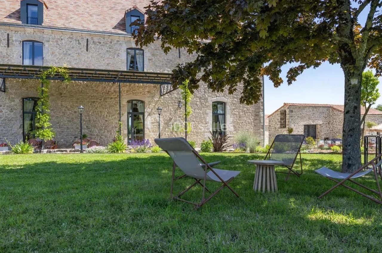 Chaises longues et table basse installées sur une pelouse ombragée devant un bâtiment en pierre avec des fenêtres.
