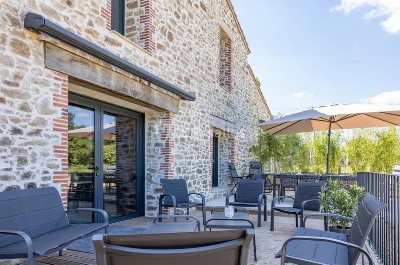 Terrasse en bois aménagée avec des fauteuils, des tables et des parasols, adossée à une maison en pierre.