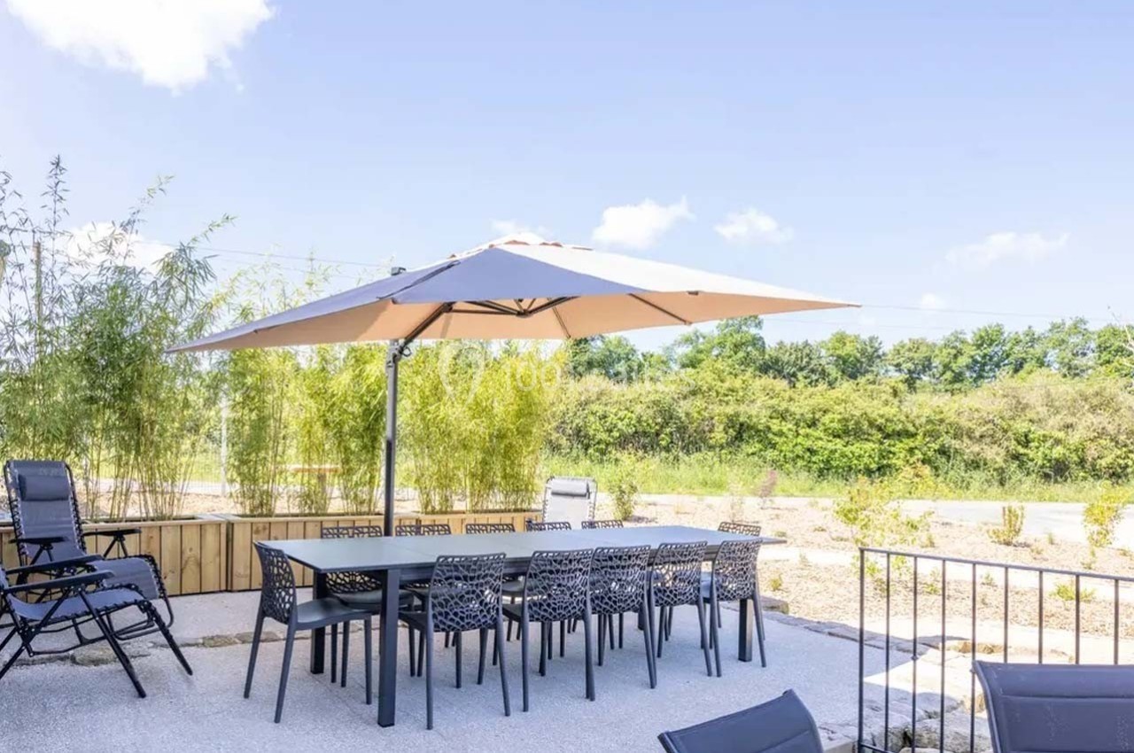 Table et chaises de jardin sous un grand parasol beige, entourées de végétation et d'un ciel dégagé.