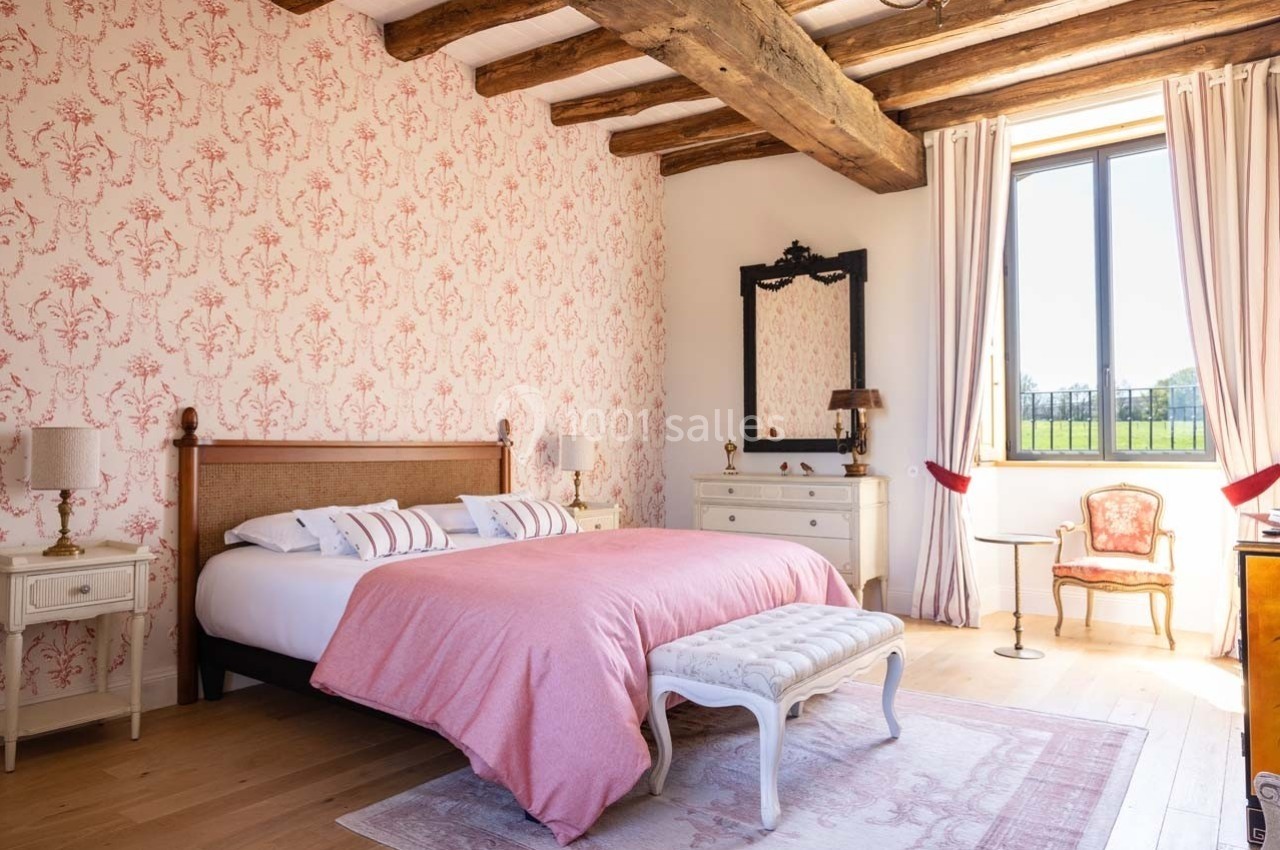 Chambre lumineuse avec lit double, couvre-lit rose, poutres apparentes, papier peint floral et vue sur un jardin.