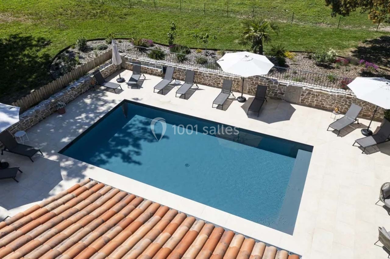 Vue aérienne d'une piscine rectangulaire entourée de chaises longues, parasols et d'un jardin verdoyant.