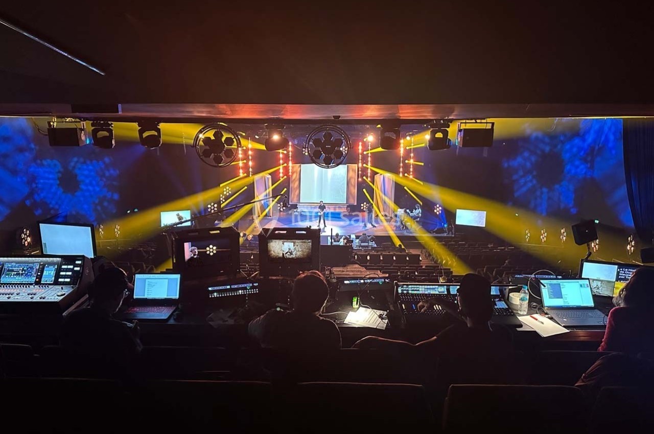 Salle de spectacle vue depuis la régie, avec techniciens et éclairages colorés dirigés vers la scène.