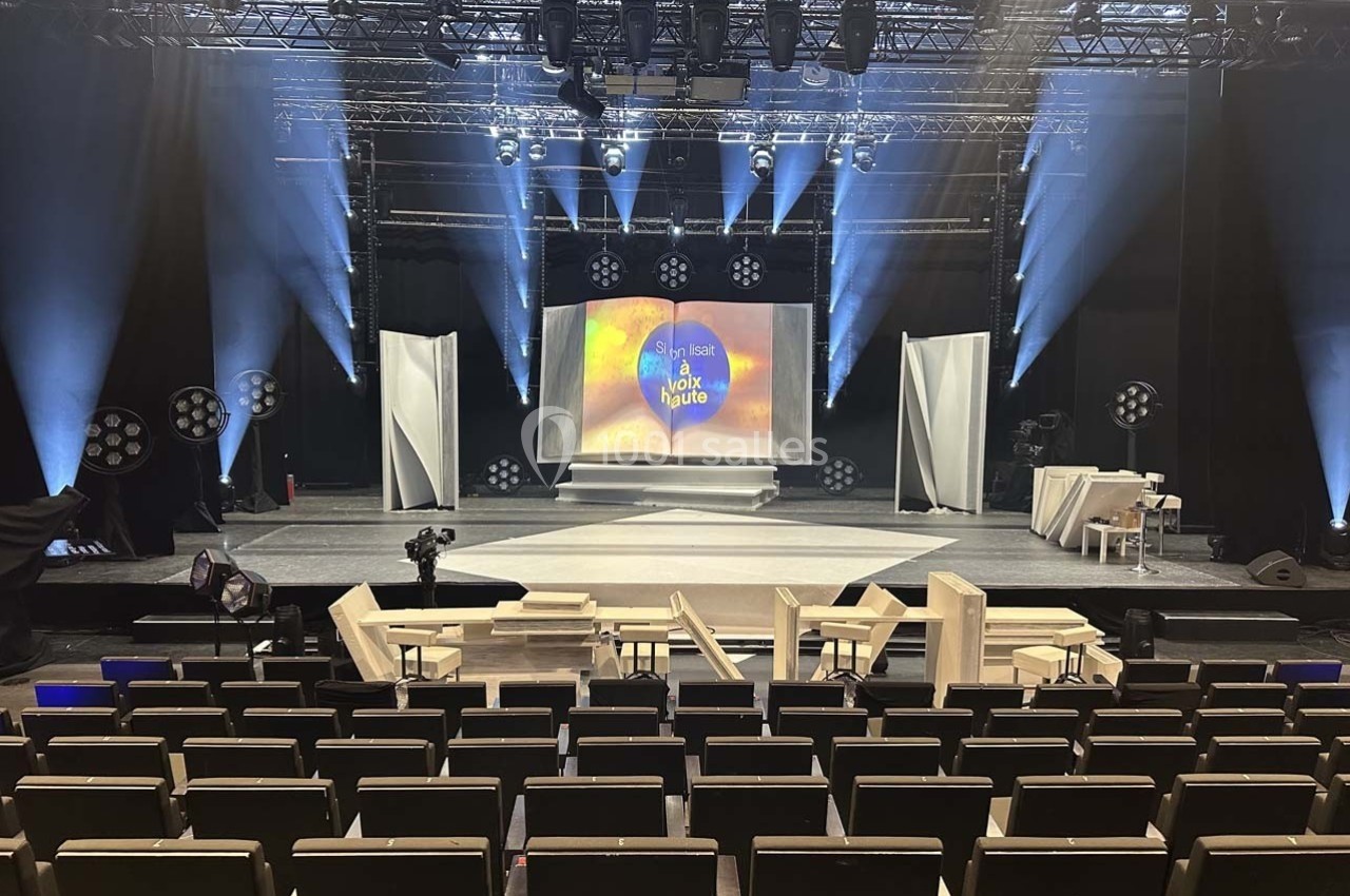 Scène de spectacle avec écran central illuminé, sièges vides au premier plan et éclairages de scène activés.