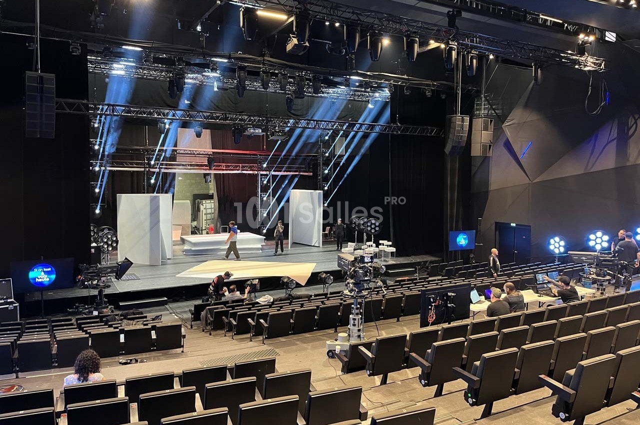 Scène de théâtre en préparation avec des techniciens et des éclairages, vue depuis les sièges d'une salle vide.