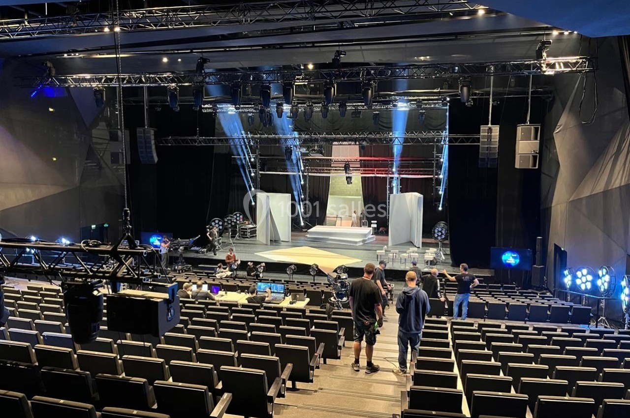 Salle de spectacle vide avec scène éclairée, techniciens en préparation et équipements techniques visibles.