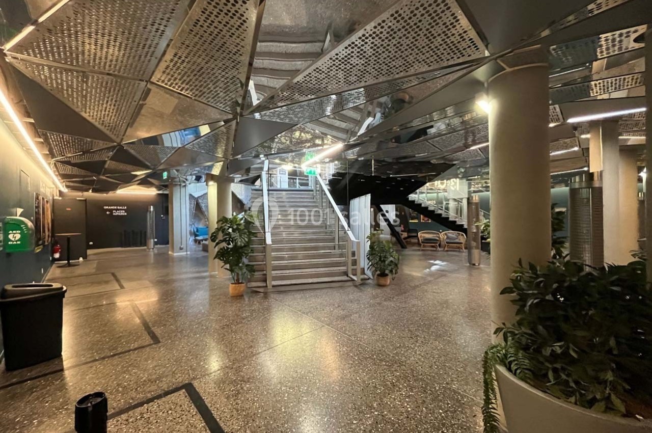Escalier central dans un hall moderne avec plafond métallique, plantes en pot et éclairage tamisé.