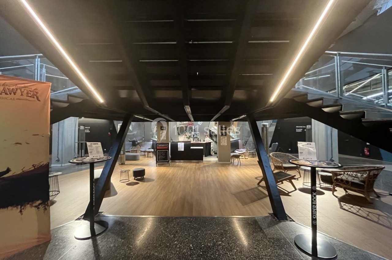 Vue d'un espace d'accueil moderne avec un escalier central, des tables hautes et des fauteuils en rotin.