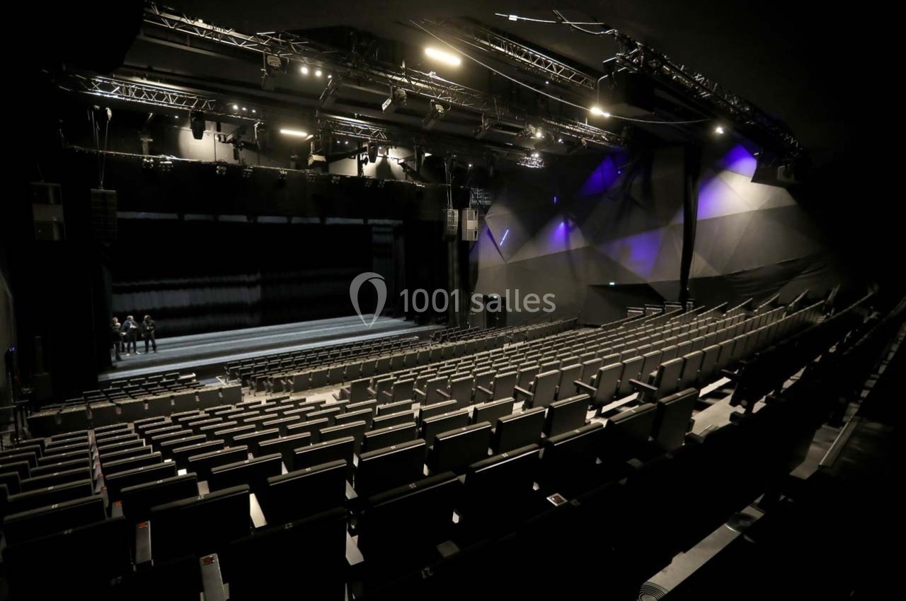 Salle de spectacle vide avec rangées de sièges, scène éclairée et projecteurs suspendus au plafond.