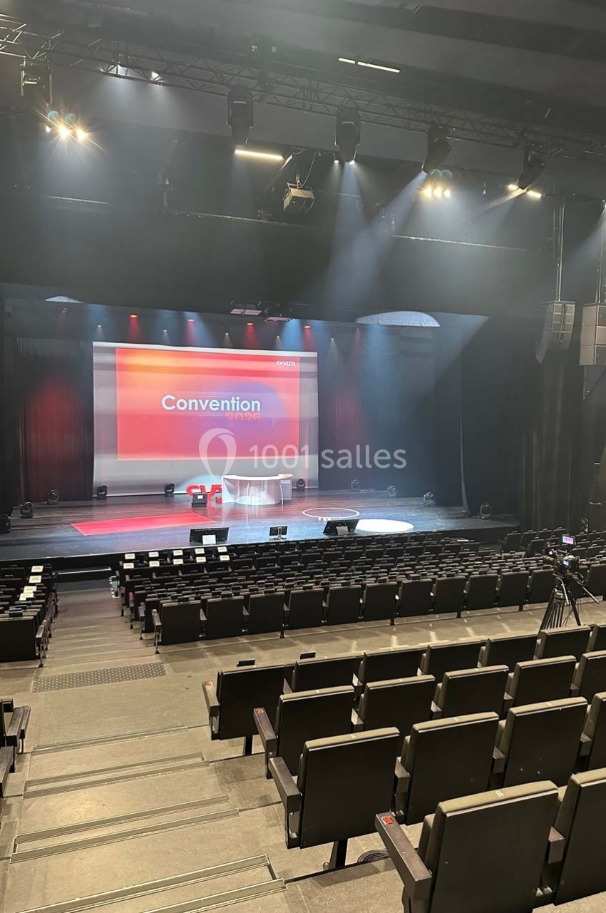 Salle de conférence avec scène éclairée, écran affichant ’Convention’ et rangées de sièges vides au premier plan.