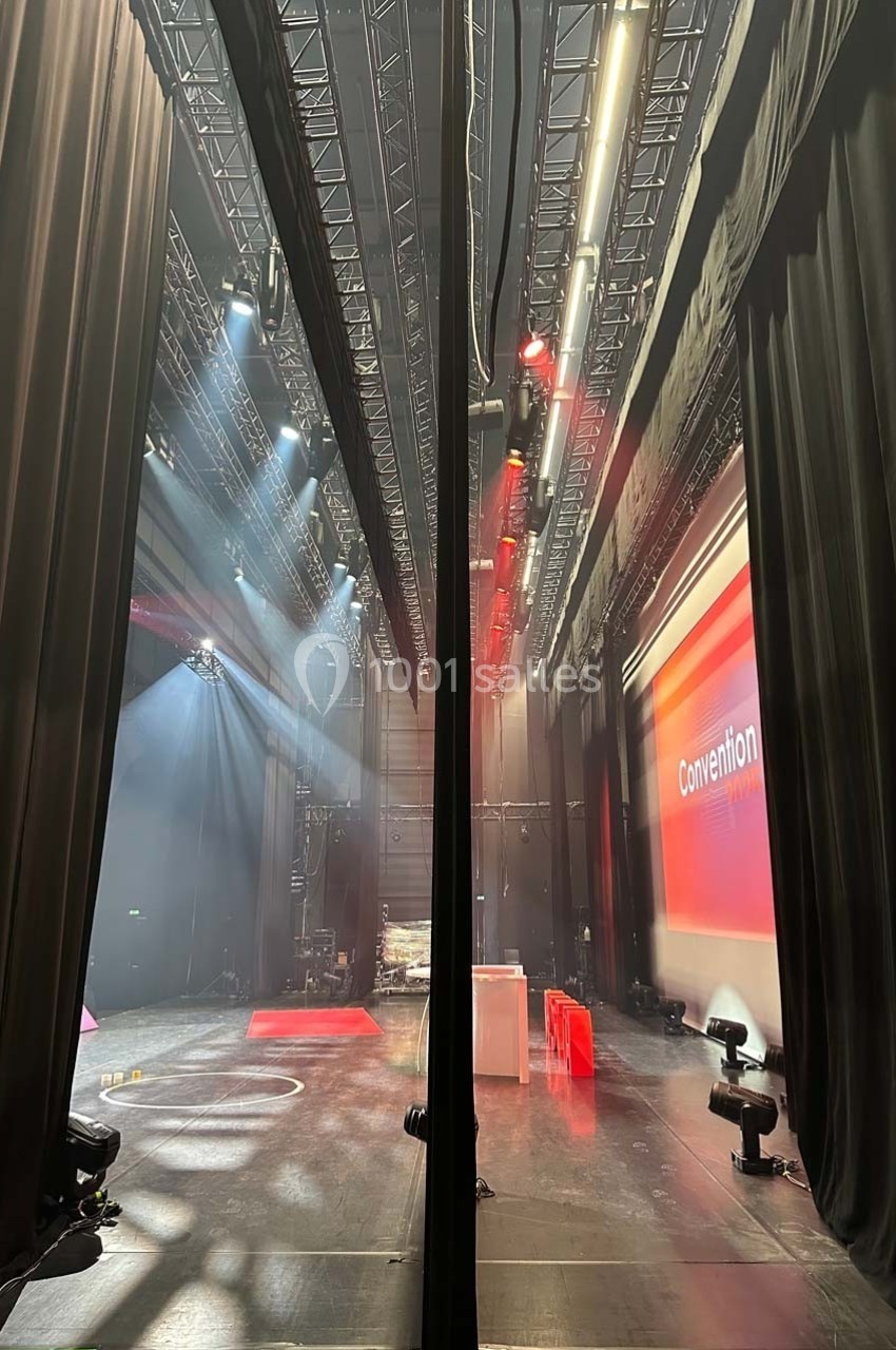 Vue des coulisses d'une scène avec rideaux noirs, éclairages de scène et écran affichant ’Convention’.