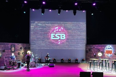 Scène éclairée avec instruments de musique, tabourets et écran affichant un logo ’ESB’ sur un fond de briques.