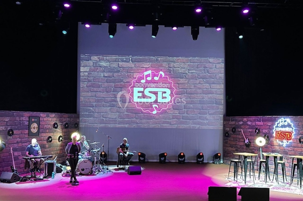 Scène éclairée avec instruments de musique, tabourets et écran affichant un logo ’ESB’ sur un fond de briques.