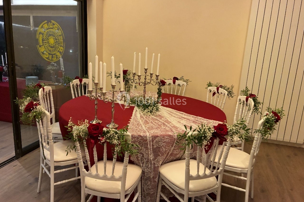 Table ronde décorée avec une nappe rouge et blanche, chandeliers, roses rouges et chaises blanches ornées de fleurs.