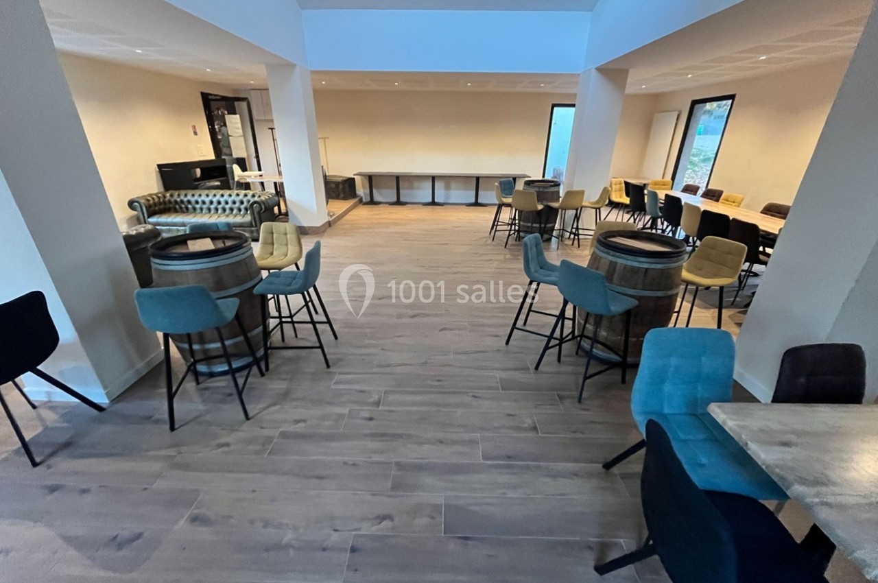 Salle lumineuse avec parquet, tables, chaises variées et tonneaux en bois utilisés comme supports.