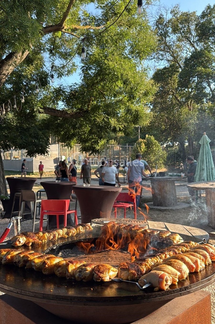Cuisson de brochettes et saucisses sur un barbecue circulaire en plein air, avec des tables et des arbres en arrière-plan.