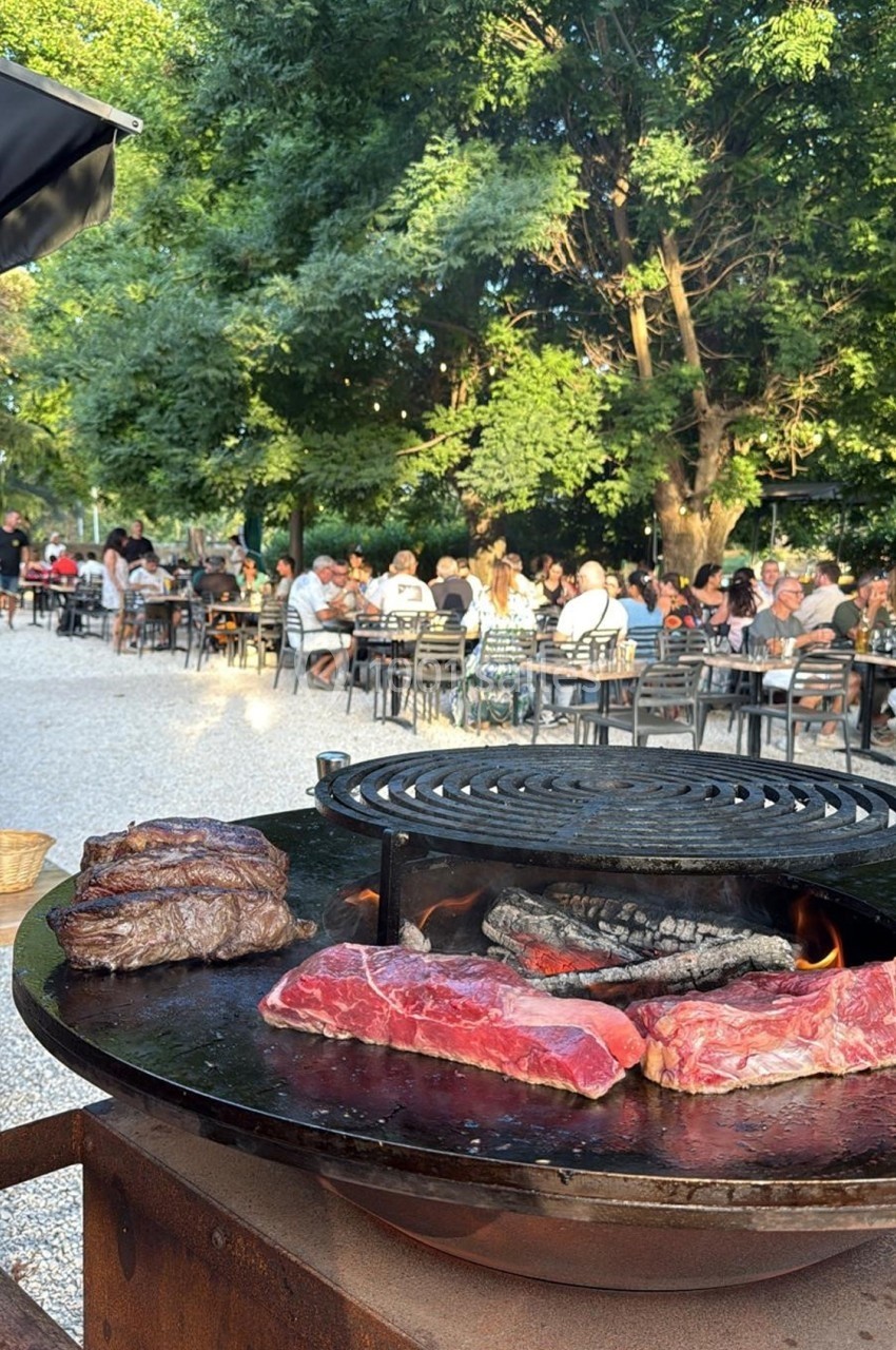 Viande grillant sur un barbecue en plein air, avec des tables occupées par des convives en arrière-plan sous des arbres.
