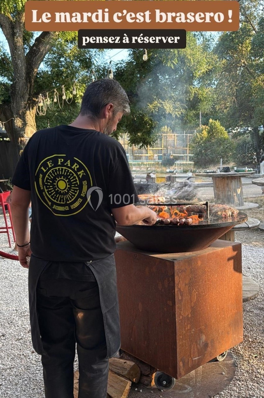 Homme cuisinant des brochettes sur un brasero en extérieur, entouré d'arbres et de mobilier de jardin.
