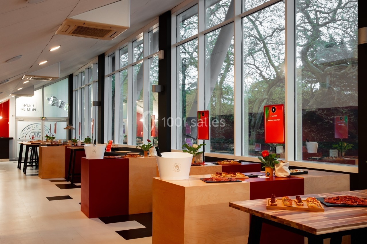 Salle lumineuse avec de grandes baies vitrées, tables en bois présentant des plats et décorations simples.