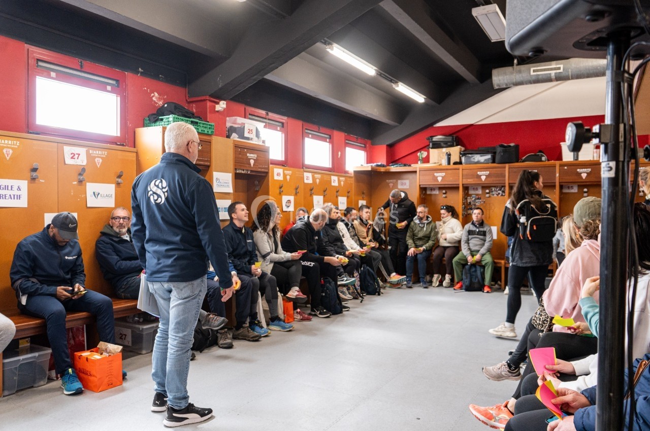 Des personnes assises dans un vestiaire discutent lors d'une réunion, entourées de casiers et d'équipements.