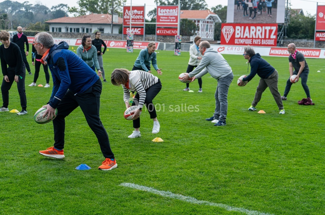 Des personnes participent à un atelier de rugby sur un terrain, tenant des ballons et effectuant des exercices en groupe.