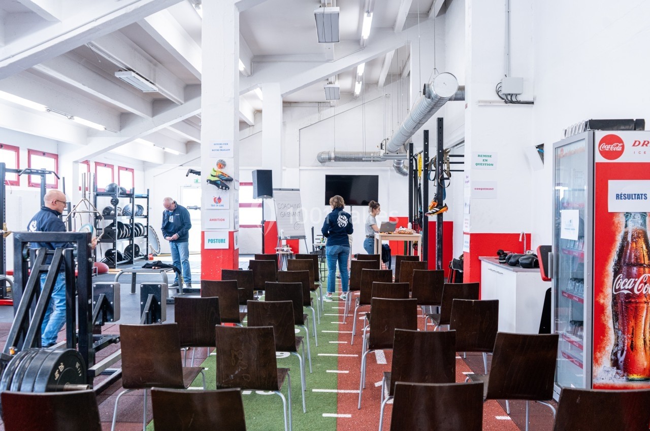 Salle de sport aménagée pour une présentation, avec des chaises alignées, un tableau blanc et des équipements sportifs.