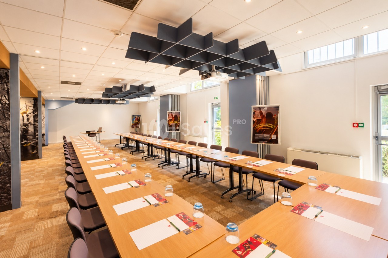 Salle de réunion avec tables en U en bois, chaises brunes, panneaux acoustiques noirs au plafond, lumière naturelle et matériel de travail disposé.