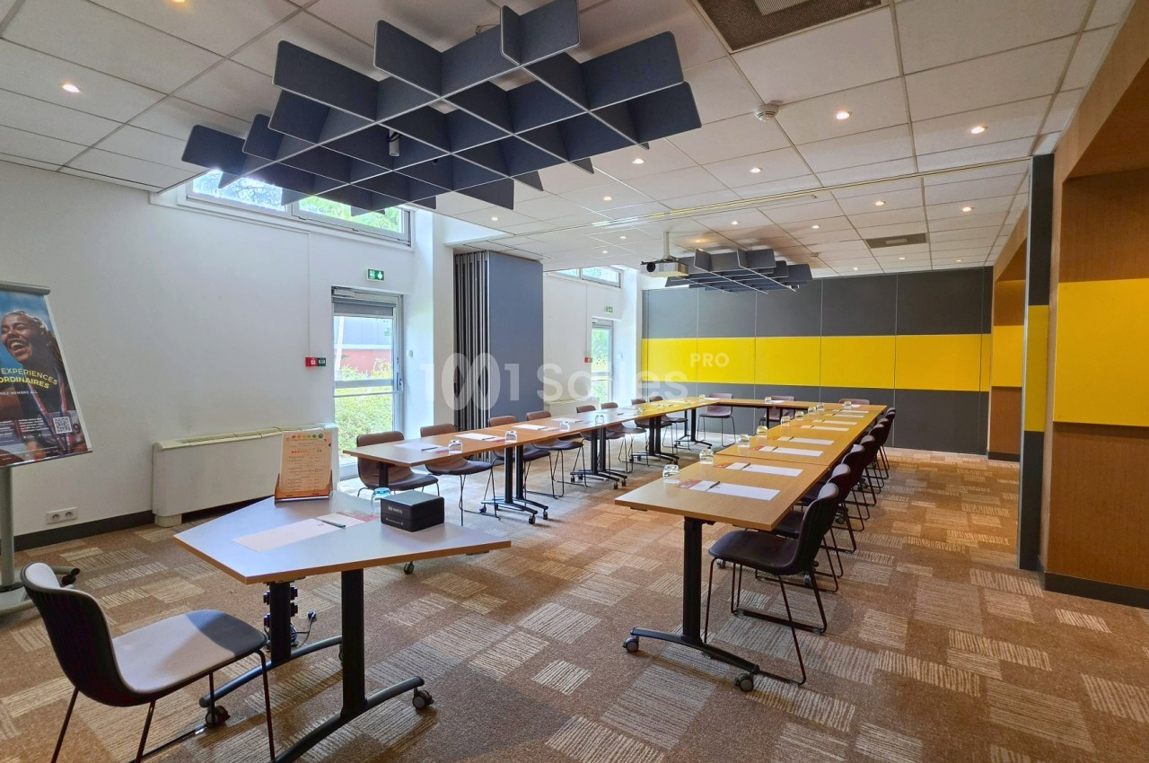Salle de réunion avec tables rectangulaires alignées, chaises noires, panneaux acoustiques bleu-noir au plafond et murs jaunes - gris.