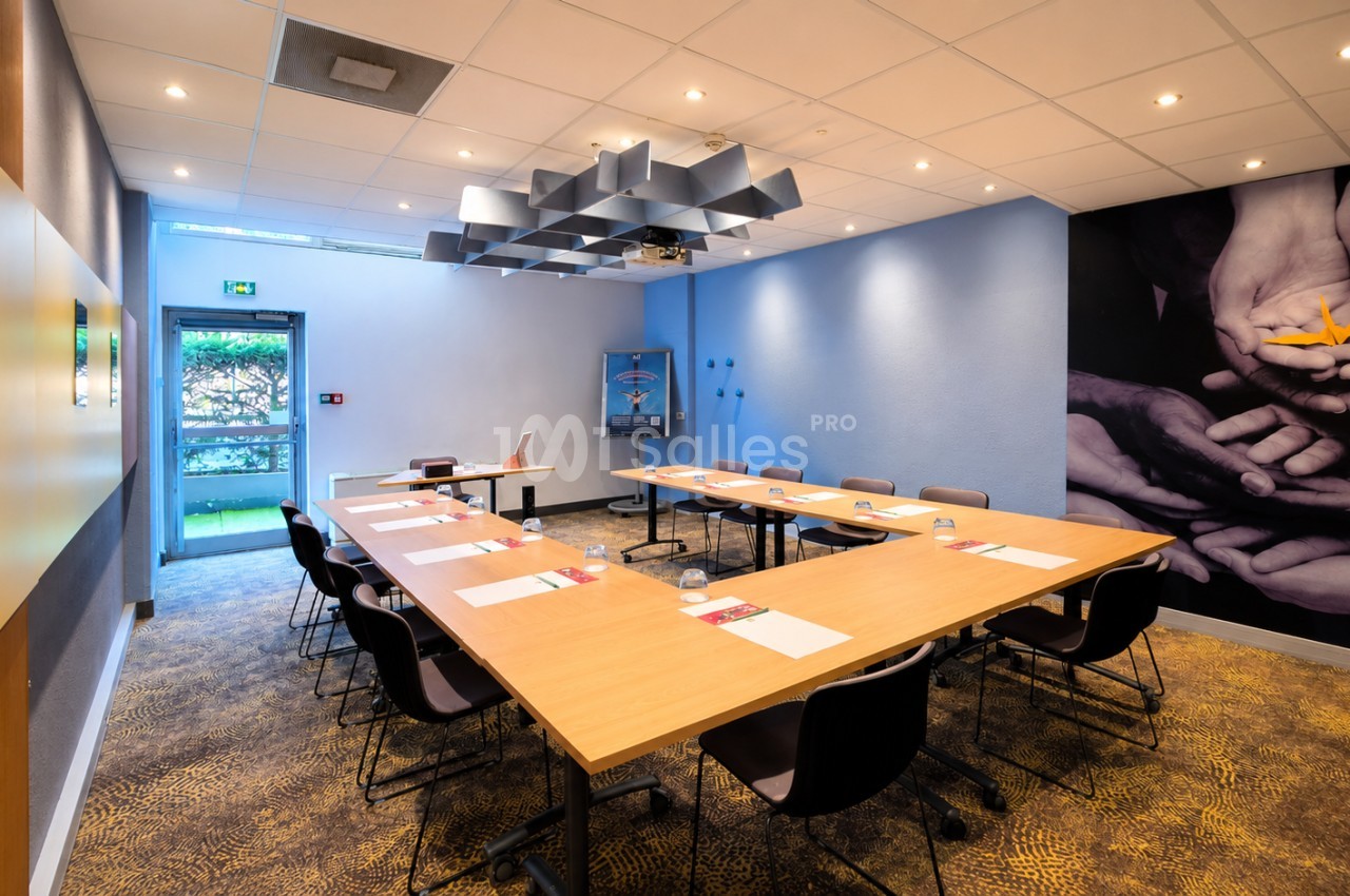 Salle de réunion rectangulaire avec tables en U, chaises noires, plafond acoustique et murs bleus.
