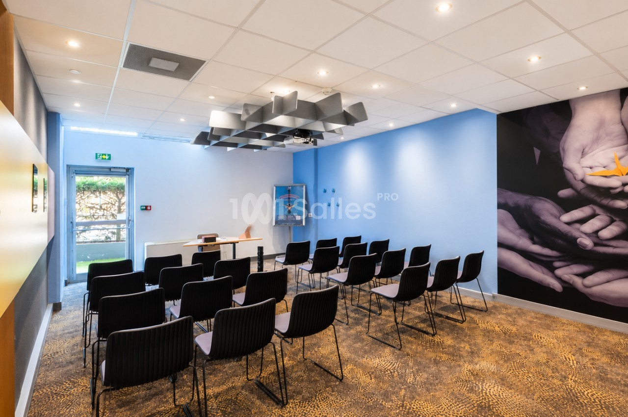 Salle de réunion avec murs bleus, chaises noires en rangées, bureau blanc et baies vitrées donnant sur l'extérieur.