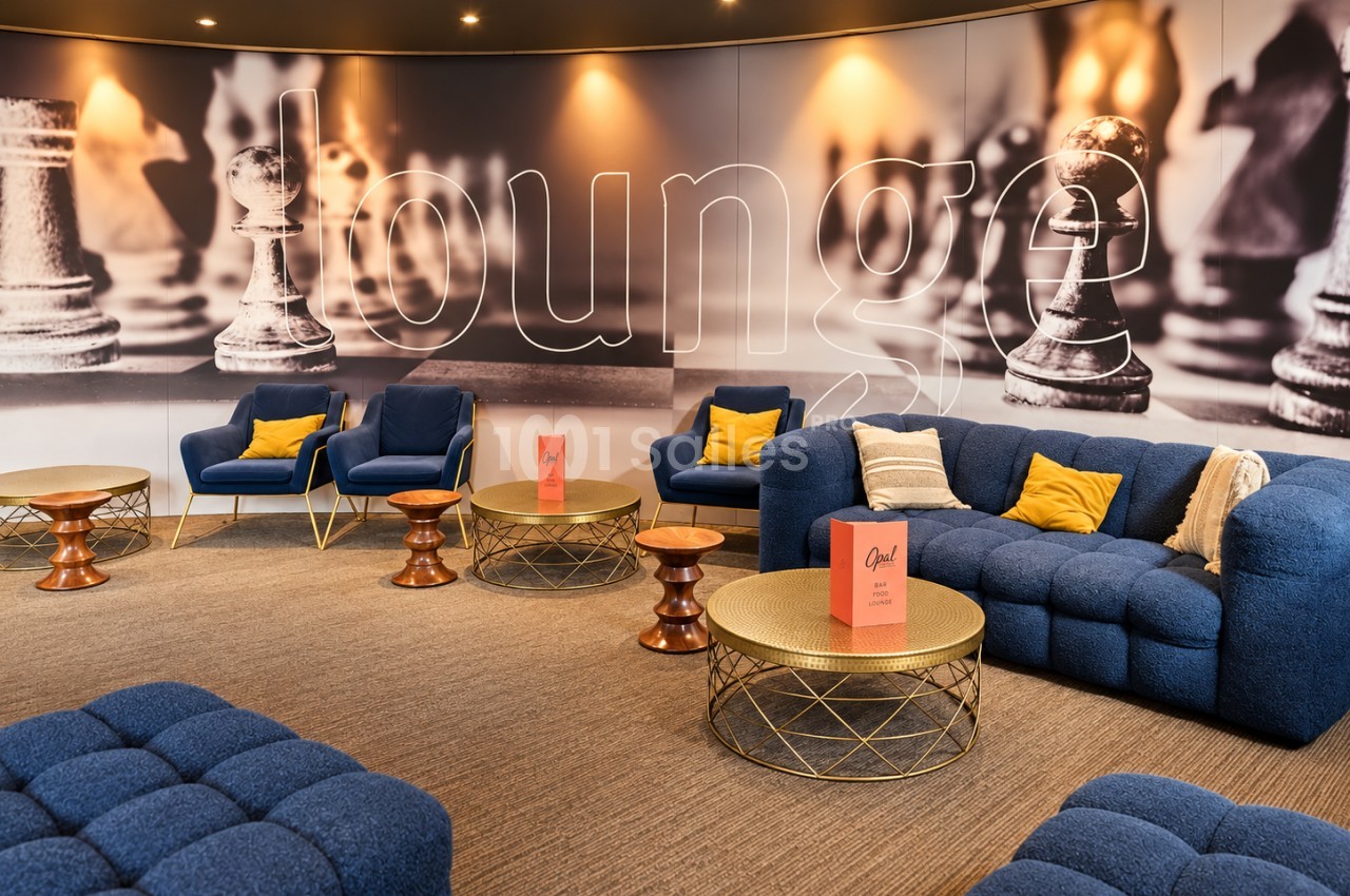 Salon lounge avec canapé et fauteuils bleu marine, tables basses dorées, mur éclairé avec motif d'échecs géants en arrière-plan.
