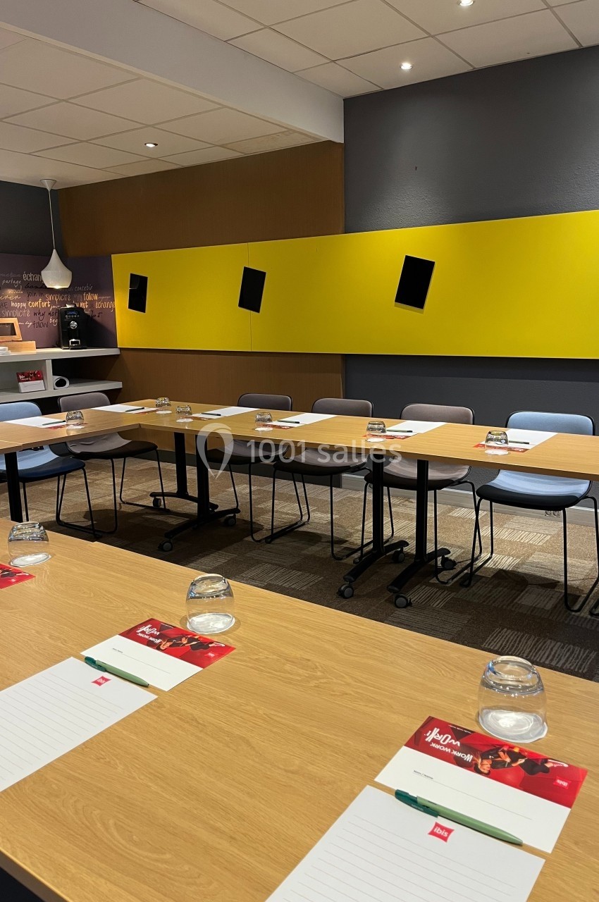 Salle de réunion moderne avec tables disposées en U, chaises, blocs-notes, verres et mur jaune en arrière-plan.
