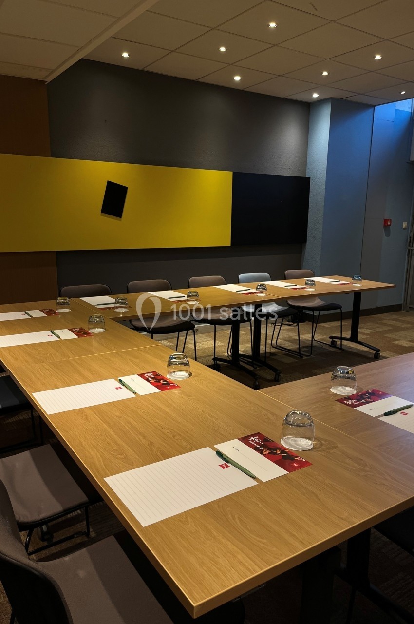 Salle de réunion avec tables en U, chaises, blocs-notes, stylos et verres d'eau, éclairée par des spots au plafond.