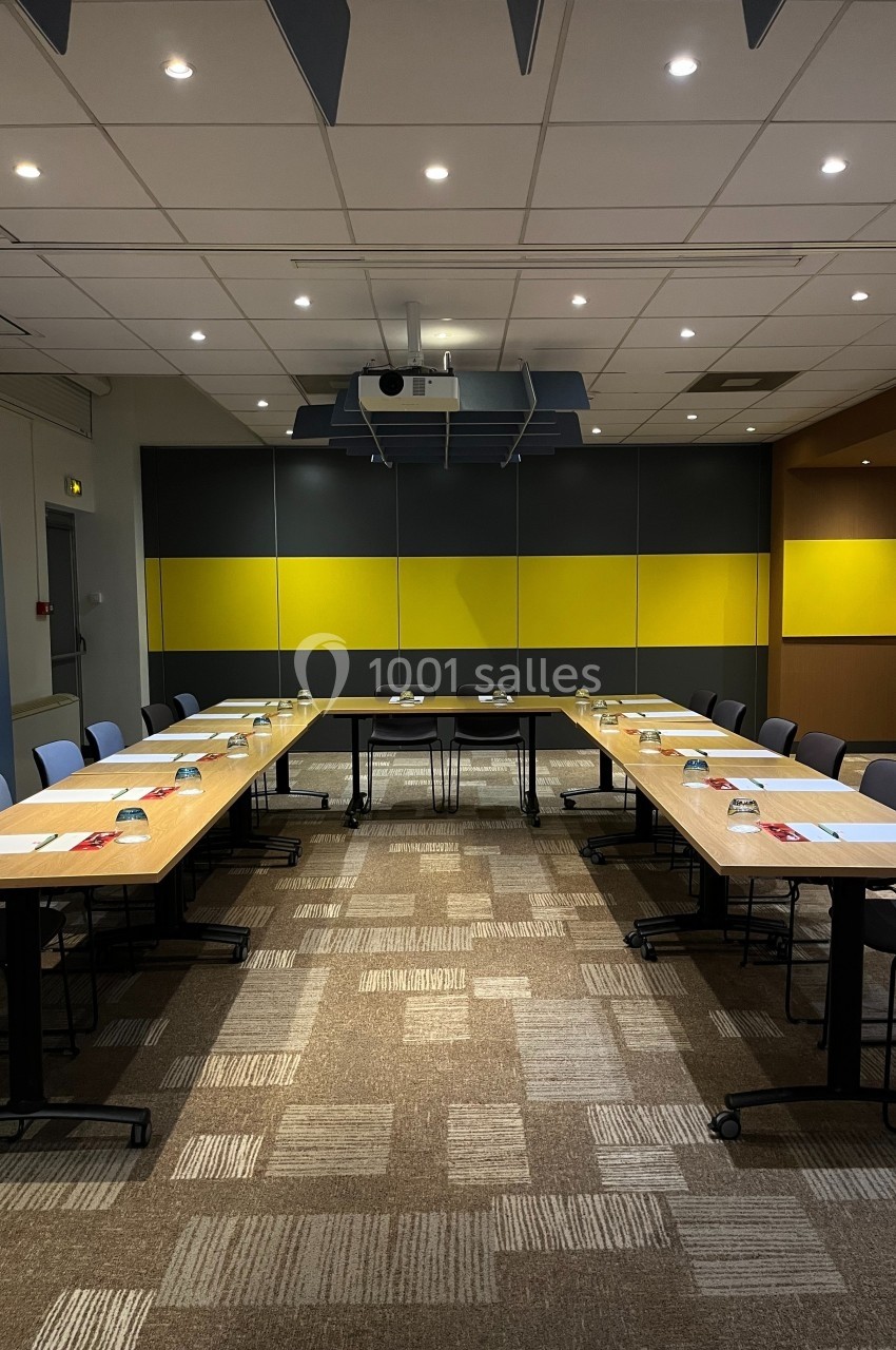 Salle de réunion moderne avec tables disposées en U, chaises alignées et projecteur au plafond.