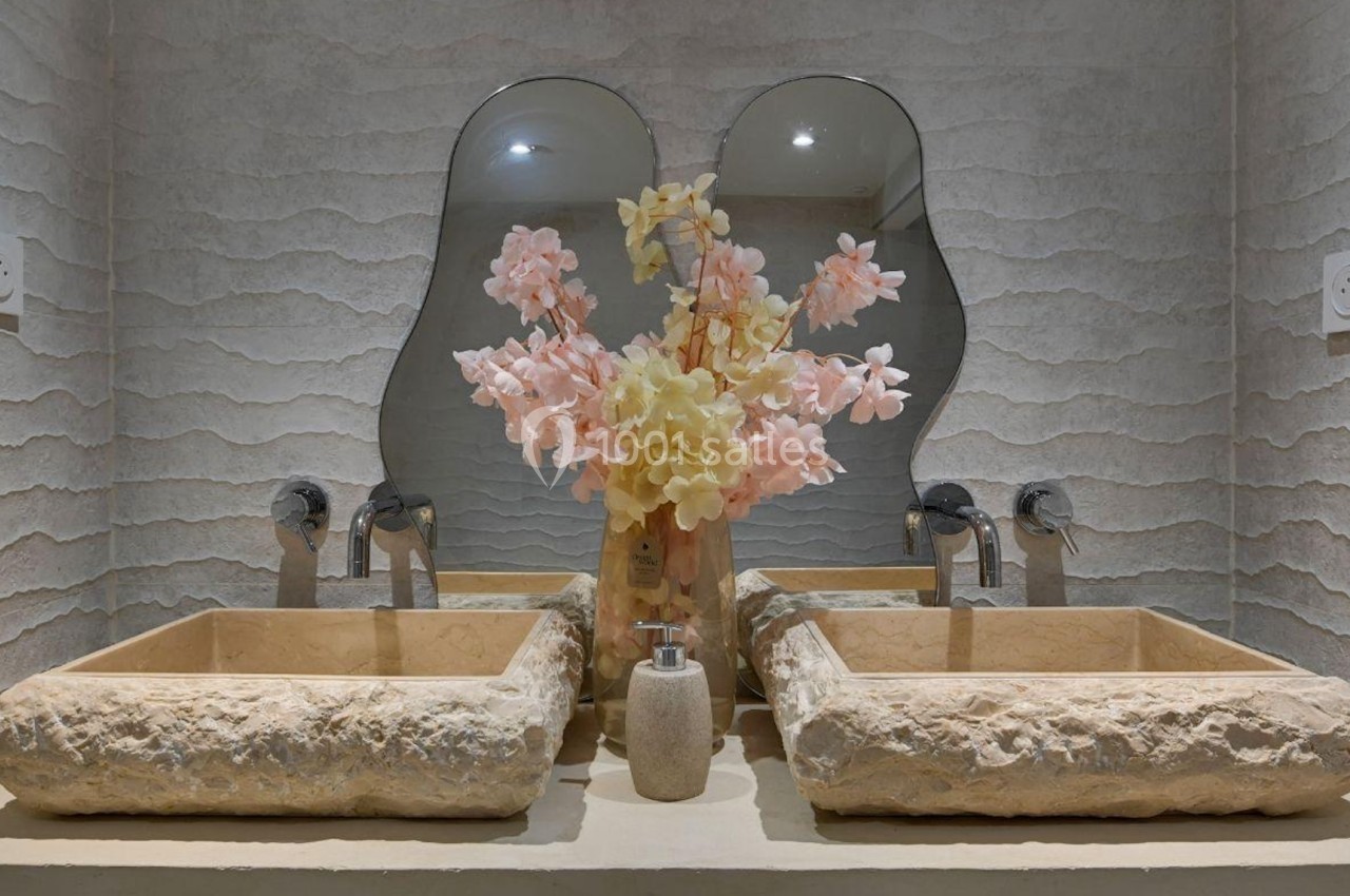 Deux lavabos en pierre avec un vase de fleurs pastel au centre, devant un miroir à double courbure.