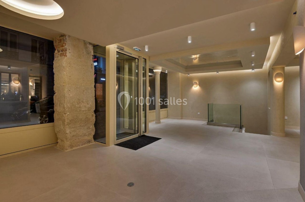 Entrée d'un bâtiment moderne avec sol en carrelage clair, murs beige, éclairage encastré et porte vitrée automatique.