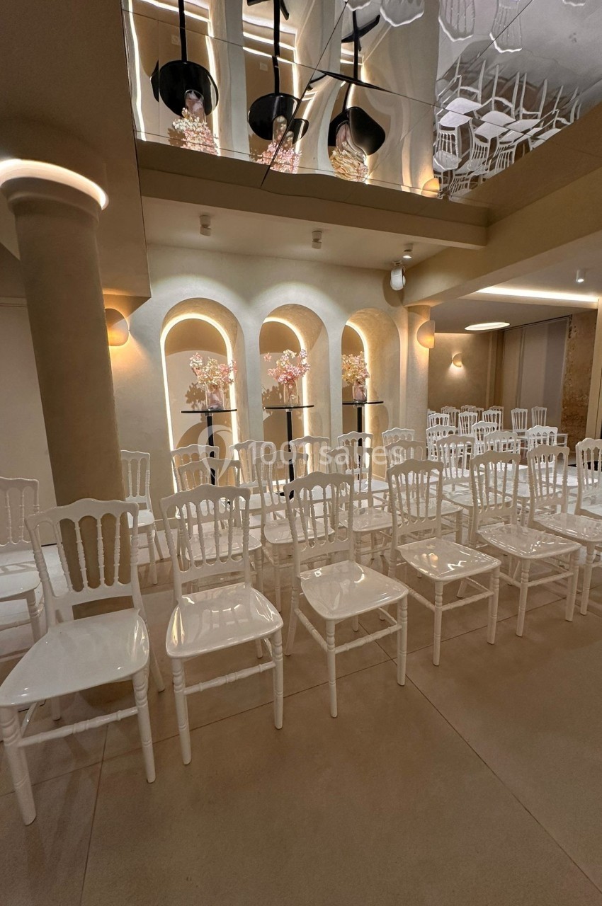 Salle décorée pour un événement avec des chaises blanches alignées, des arches lumineuses et des compositions florales.