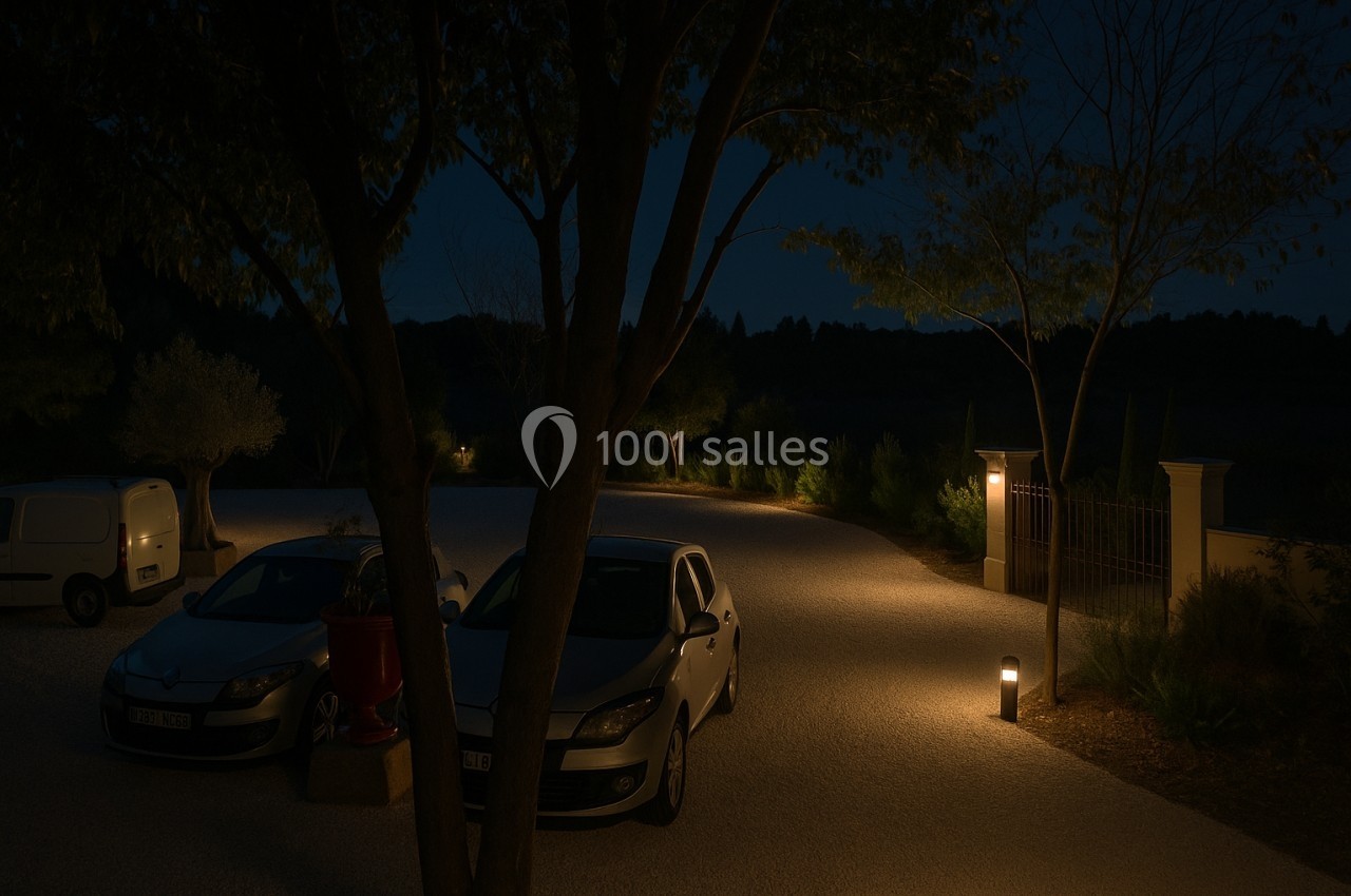 Allée gravillonnée éclairée la nuit, bordée d'arbres et de lampadaires, avec des voitures stationnées.