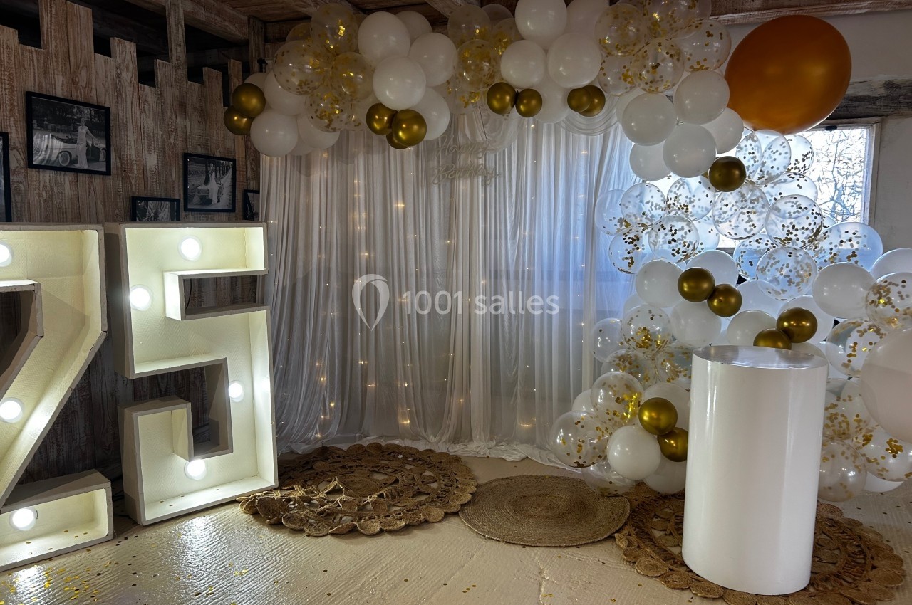 Décoration festive avec ballons blancs et dorés, rideau lumineux, chiffres lumineux ’25’ et socle blanc.