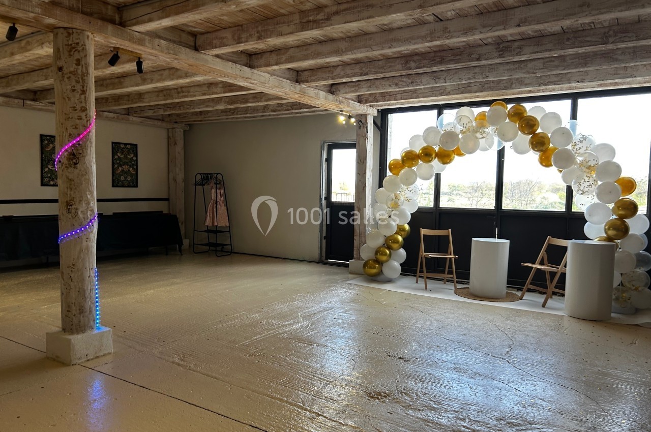 Salle lumineuse avec poutres apparentes, arche de ballons blancs et dorés, et chaises disposées près d'une fenêtre.