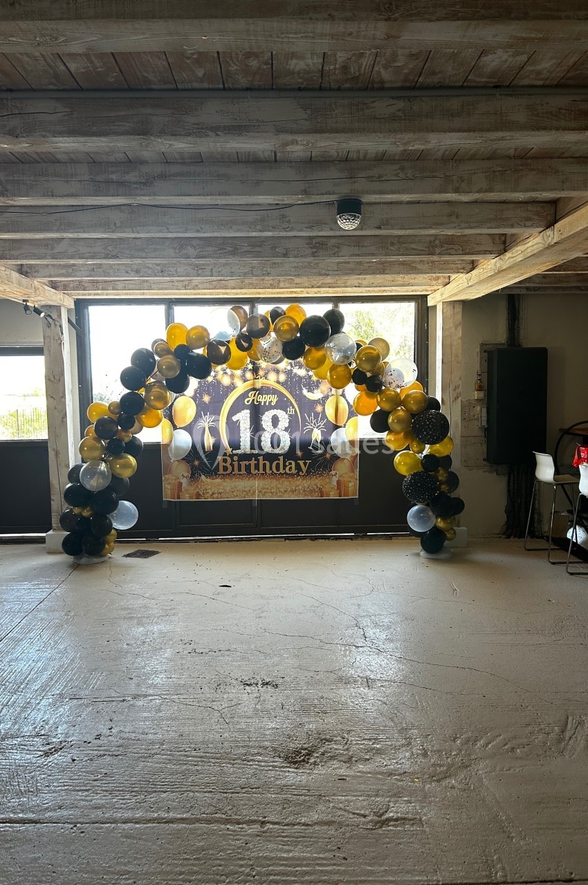 Arche de ballons noirs, dorés et argentés devant une bannière ’Happy 18th Birthday’ dans un espace en bois.