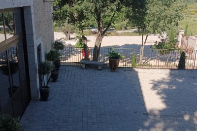 Pot rouge en céramique posé sur un socle en pierre, entouré d'arbres et de gravier dans une cour clôturée.
