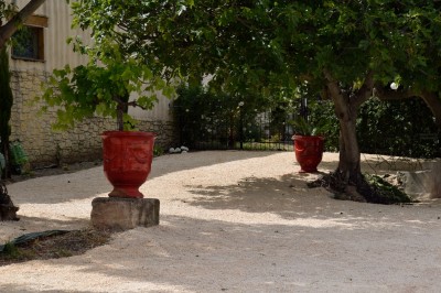 Pot rouge en céramique posé sur un socle en pierre, entouré d'arbres et de gravier dans une cour clôturée.