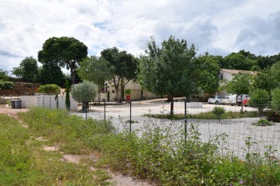 Pot rouge en céramique posé sur un socle en pierre, entouré d'arbres et de gravier dans une cour clôturée.