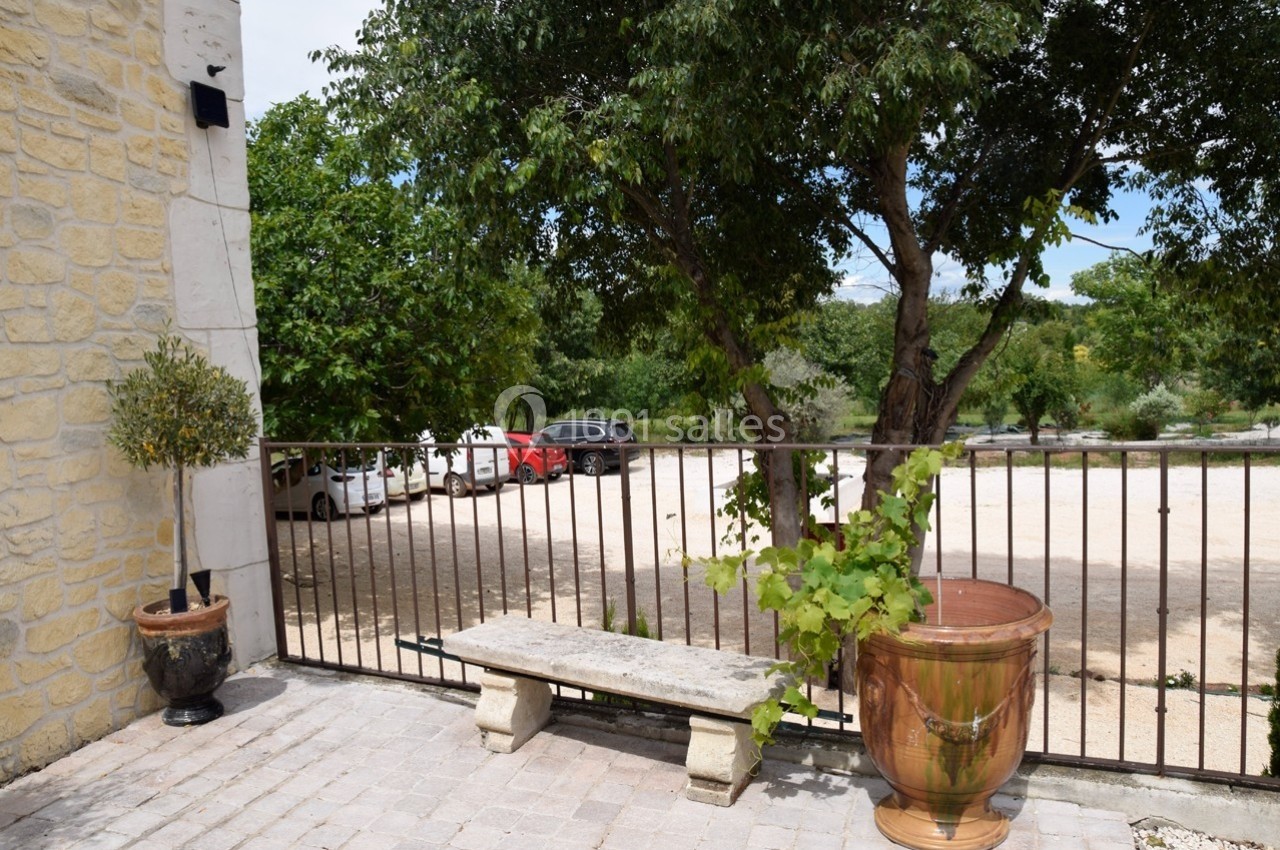 Terrasse en pierre avec un banc, une plante en pot et une vue sur un parking bordé d'arbres.