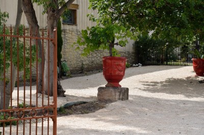 Pot rouge en céramique posé sur un socle en pierre, entouré d'arbres et de gravier dans une cour clôturée.