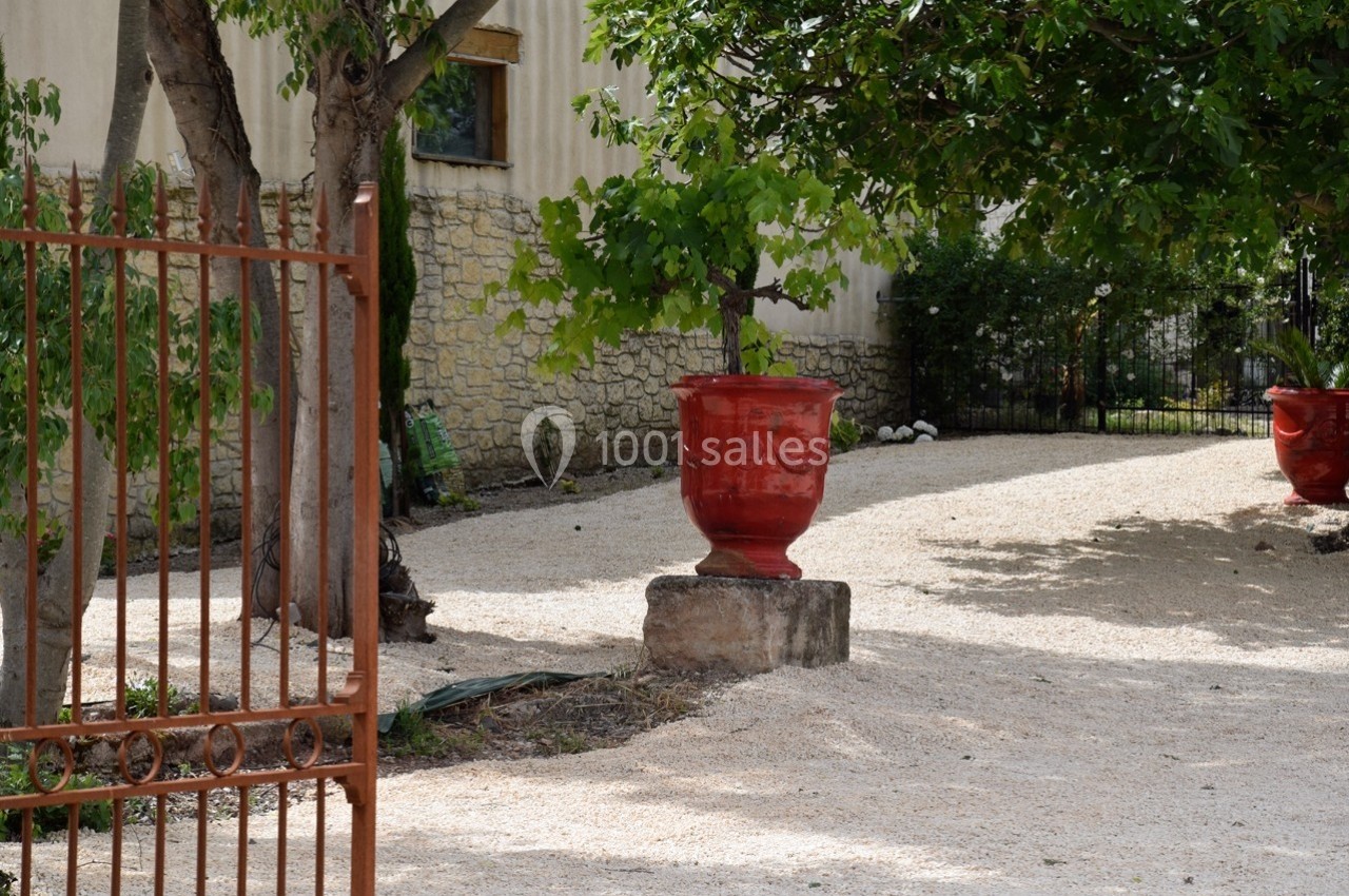 Pot rouge en céramique posé sur un socle en pierre, entouré d'arbres et de gravier dans une cour clôturée.