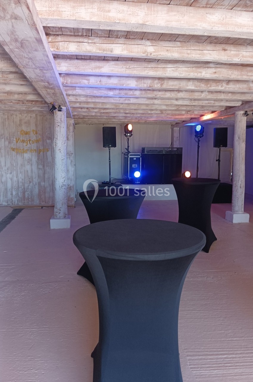 Salle avec tables hautes noires, éclairage coloré et équipement audio installé dans un espace à poutres apparentes.