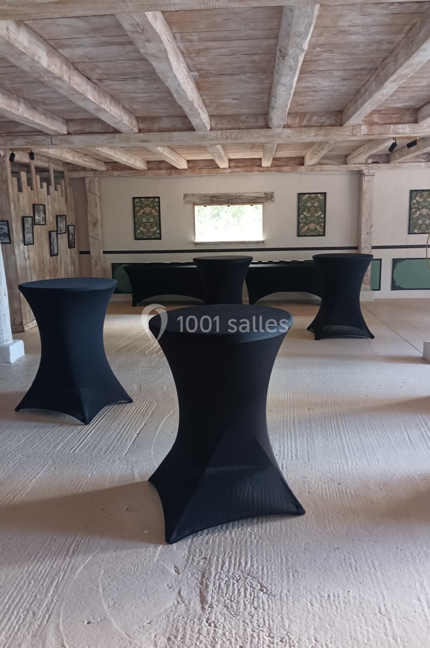 Salle avec des tables hautes noires habillées de housses, poutres apparentes et décorations murales.