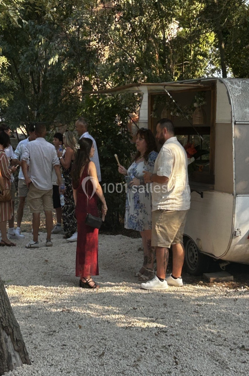 Un groupe de personnes discute près d'une caravane blanche dans un espace extérieur ombragé.